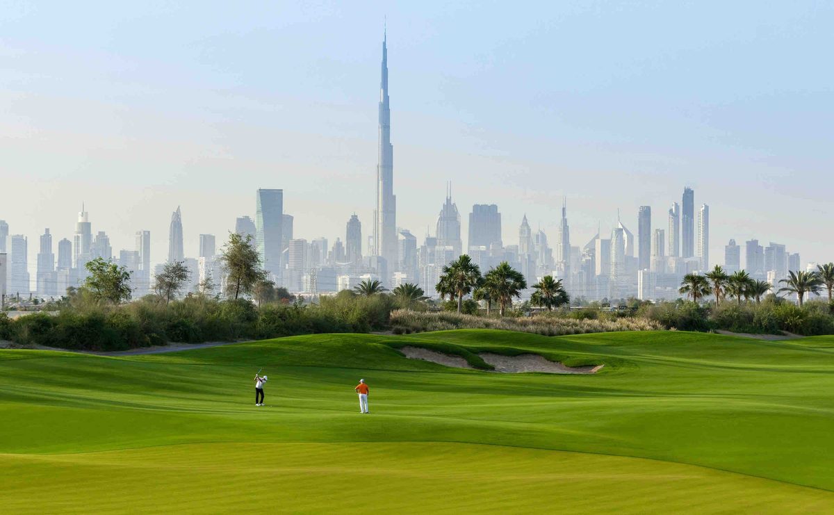 The Dubai Hills golf course with two golfers on the fairway, palms beyond it and the Downtown Dubai skyline in the haze