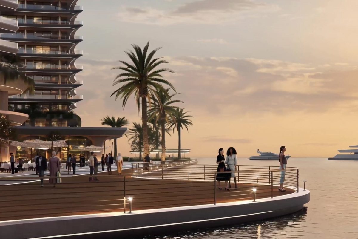 The marina promenade at dusk: a curved timber deck along the water, palms, a residential tower above the retail colonnade and yachts moored beyond