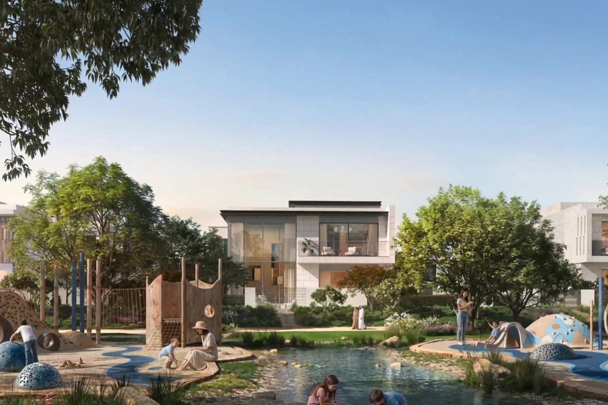 A neighbourhood park with a shallow water-play channel, timber play structures and families under mature trees, a villa behind